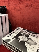 A stack of black and white magazines is placed on a textured red surface. The top magazine features a cover with Japanese text and a retro-styled illustration or photograph of a smiling person wearing a hat, possibly a caricature, with another figure in the background.