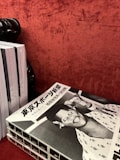 A stack of black and white magazines is placed on a textured red surface. The top magazine features a cover with Japanese text and a retro-styled illustration or photograph of a smiling person wearing a hat, possibly a caricature, with another figure in the background.