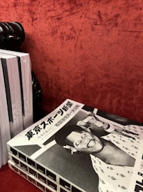 A stack of black and white magazines is placed on a textured red surface. The top magazine features a cover with Japanese text and a retro-styled illustration or photograph of a smiling person wearing a hat, possibly a caricature, with another figure in the background.