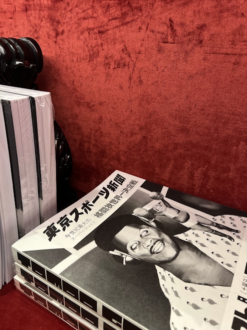 A stack of black and white magazines is placed on a textured red surface. The top magazine features a cover with Japanese text and a retro-styled illustration or photograph of a smiling person wearing a hat, possibly a caricature, with another figure in the background.