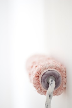 A paint roller covered in pink paint is pressing against a white wall. The roller appears to be in mid-action, with the paint on the roller appearing thick and textured.