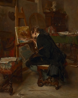 A person seated in an art studio, deeply focused on painting a canvas placed on an easel. The room is dimly lit, adding a warm, contemplative atmosphere. Surrounding the artist are various artistic tools and furniture, including a decorative stool, a table with papers, and a wooden cabinet. A bust sculpture is visible in the background, suggesting an inspirational setting.