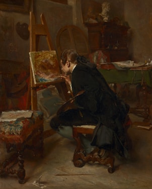 A person seated in an art studio, deeply focused on painting a canvas placed on an easel. The room is dimly lit, adding a warm, contemplative atmosphere. Surrounding the artist are various artistic tools and furniture, including a decorative stool, a table with papers, and a wooden cabinet. A bust sculpture is visible in the background, suggesting an inspirational setting.