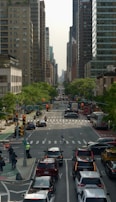 An urban street scene with multiple lanes of traffic, tall buildings on either side, and green trees lining the sidewalks. Vehicles are stopped at a traffic light, and a few pedestrians can be seen crossing the street. Bicycles are parked on the left, and a traffic signal and bus are visible.
