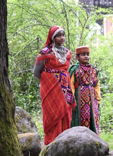 Two individuals are dressed in vibrant, traditional garments adorned with intricate patterns and colorful designs. They are standing amidst a lush green backdrop with moss-covered rocks in a natural setting. The woman is wearing ornate silver jewelry, including a headpiece and layered necklaces, adding to the cultural richness.