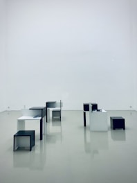Several minimalist, geometric tables and stools in black and white are arranged in a spacious, empty gallery with a glossy, reflective floor.