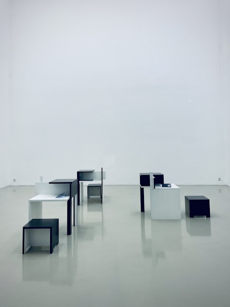 Several minimalist, geometric tables and stools in black and white are arranged in a spacious, empty gallery with a glossy, reflective floor.