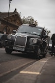 A black London taxi with distinct vintage design details is parked on a street. The background includes a historic building with architectural elements visible. The sky appears overcast, contributing to a subdued ambiance.