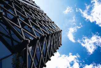 A modern architectural structure featuring a lattice design of metallic rods ascending vertically against a bright blue sky with scattered white clouds.