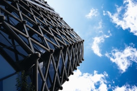 A modern architectural structure featuring a lattice design of metallic rods ascending vertically against a bright blue sky with scattered white clouds.