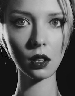 A black-and-white close-up portrait of a person with light hair, large eyes with dark eyeliner, and glossy lipstick. A few strands of hair fall across the face, and the lighting highlights the facial features while creating shadows.