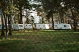 A row of white caravans is parked among tall pine trees in a forested area. The ground is covered with green grass, and soft sunlight filters through the trees, casting dappled shadows. A wire mesh fence runs across the foreground.