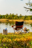 A camping chair is set up by the tranquil lake, next to a white cooler and a stack of firewood on the grass. Trees and vegetation surround the lake, creating a serene outdoor setting.