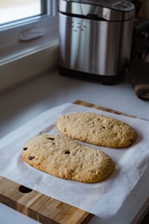 Two freshly baked loaves of bread with embedded pieces of fruit sit on a parchment-lined wooden cutting board. The scene is set in a kitchen with a bread machine visible in the background, alongside a window that allows natural light to illuminate the setup.