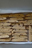 A tray of freshly baked biscotti arranged in rows on parchment paper. The biscotti are golden brown with visible pieces of dried fruit and nuts.