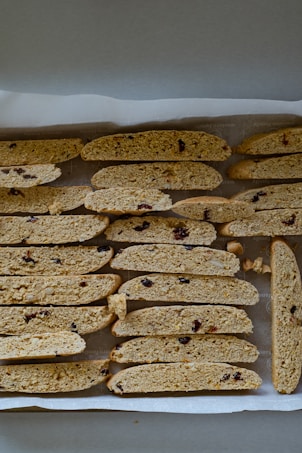 A tray of freshly baked biscotti arranged in rows on parchment paper. The biscotti are golden brown with visible pieces of dried fruit and nuts.
