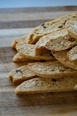 A stack of sliced biscotti placed on a wooden cutting board. The biscotti have a golden-brown color with visible nuts and dried fruit embedded within. The texture appears crunchy and crumbly.