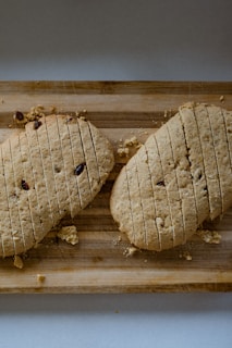 Two baked loaves of bread scored with parallel cuts lie on a wooden cutting board with crumbs scattered around. The loaves have a rustic appearance with bits of dried fruit embedded in them.
