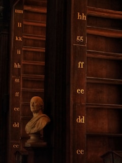A classical bust sculpture placed in front of a large, dark wooden bookcase with rows of shelves labeled with alphabetic letters. The lighting is dim, creating a warm and historical atmosphere.