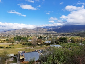 A sprawling valley with scattered houses and trees under a partly cloudy sky. Rolling hills and mountains form the backdrop, with a variety of green foliage in the foreground.