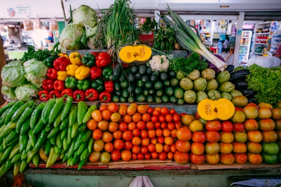 A vibrant display of fresh vegetables on a market stall, featuring a variety of colors and types. The assortment includes green peppers, red tomatoes, cucumbers, squash, lettuce, and scallions. The arrangement is neat and plentiful, creating an inviting and abundant appearance. Background elements suggest a bustling market environment.