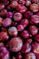 A close-up view of a large pile of red onions. The onions vary in size and have a shiny, slightly rough surface texture with a mix of purple and red hues. Some onions have dry, papery skins partially peeling off.