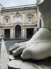 A large marble foot sculpture is prominently displayed in the foreground, highlighting its intricate detailing and smooth texture. In the background, an architectural facade with columns and arches showcases classical design elements. The building features decorative carvings and inscriptions above the main entrance.