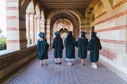 A group of five individuals wearing graduation gowns and caps walk together through a corridor with brick and stone archways. The floor is paved with patterned bricks, and the corridor is illuminated by natural light.