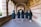 A group of five individuals wearing graduation gowns and caps walk together through a corridor with brick and stone archways. The floor is paved with patterned bricks, and the corridor is illuminated by natural light.