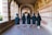 A group of five individuals wearing graduation gowns and caps walk together through a corridor with brick and stone archways. The floor is paved with patterned bricks, and the corridor is illuminated by natural light.