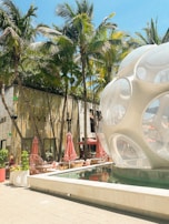 Palm trees line a luxurious outdoor shopping area featuring a modern, spherical glass structure next to a shallow reflecting pool. Red umbrellas and seating areas provide a shaded space for relaxation. The backdrop includes a high-end retail store with a sleek, contemporary design.