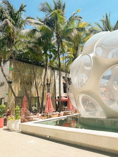 Palm trees line a luxurious outdoor shopping area featuring a modern, spherical glass structure next to a shallow reflecting pool. Red umbrellas and seating areas provide a shaded space for relaxation. The backdrop includes a high-end retail store with a sleek, contemporary design.