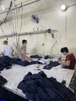Several people are working at a large table in a textile factory, engaged in folding or sewing fabric. They are surrounded by piles of dark material. The atmosphere is utilitarian with exposed pipes and wires on the walls.