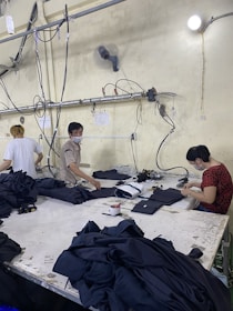 Several people are working at a large table in a textile factory, engaged in folding or sewing fabric. They are surrounded by piles of dark material. The atmosphere is utilitarian with exposed pipes and wires on the walls.