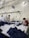 Several people are working at a large table in a textile factory, engaged in folding or sewing fabric. They are surrounded by piles of dark material. The atmosphere is utilitarian with exposed pipes and wires on the walls.