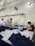Several people are working at a large table in a textile factory, engaged in folding or sewing fabric. They are surrounded by piles of dark material. The atmosphere is utilitarian with exposed pipes and wires on the walls.