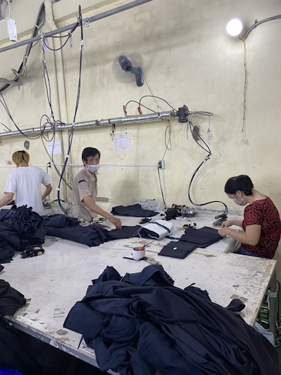 Several people are working at a large table in a textile factory, engaged in folding or sewing fabric. They are surrounded by piles of dark material. The atmosphere is utilitarian with exposed pipes and wires on the walls.