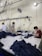 Several people are working at a large table in a textile factory, engaged in folding or sewing fabric. They are surrounded by piles of dark material. The atmosphere is utilitarian with exposed pipes and wires on the walls.