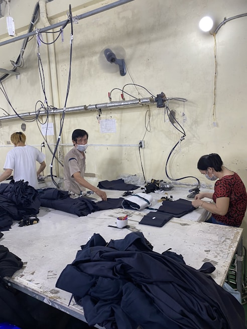 Several people are working at a large table in a textile factory, engaged in folding or sewing fabric. They are surrounded by piles of dark material. The atmosphere is utilitarian with exposed pipes and wires on the walls.