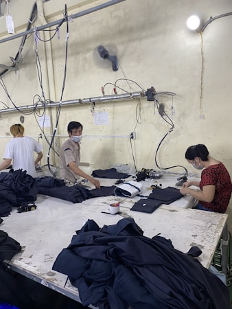Several people are working at a large table in a textile factory, engaged in folding or sewing fabric. They are surrounded by piles of dark material. The atmosphere is utilitarian with exposed pipes and wires on the walls.