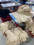 A room with sewing equipment and fabric bundles is depicted. There is a sewing machine on a table surrounded by stacks of beige fabric. Some fabric pieces are piled onto the table, while others are neatly folded in a red basket.