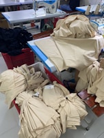 A room with sewing equipment and fabric bundles is depicted. There is a sewing machine on a table surrounded by stacks of beige fabric. Some fabric pieces are piled onto the table, while others are neatly folded in a red basket.