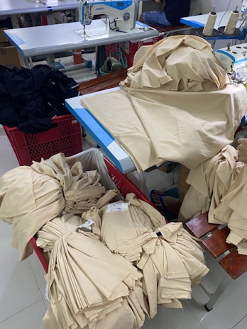 A room with sewing equipment and fabric bundles is depicted. There is a sewing machine on a table surrounded by stacks of beige fabric. Some fabric pieces are piled onto the table, while others are neatly folded in a red basket.