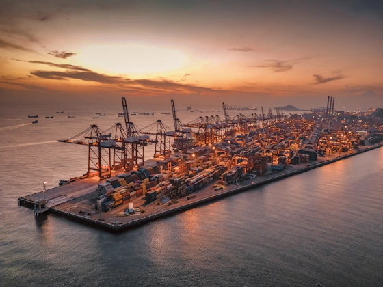 A panoramic view of a bustling Southeast Asian port with cargo ships, oil rigs, and tourist boats under a vibrant sunset.