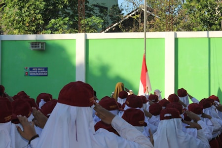 A group of school children wearing red hats and white uniforms are standing in front of a green wall, saluting towards an Indonesian flag. Trees are visible above the wall, and a sign in the background displays the message about not littering.