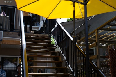 A metal staircase with a black railing leads up to a balcony. A large yellow umbrella provides shade over part of the stairs. The background reveals a house with windows and some wooden lattice structures. Partial figures of people are visible at the top of the stairs and in the shaded area below.