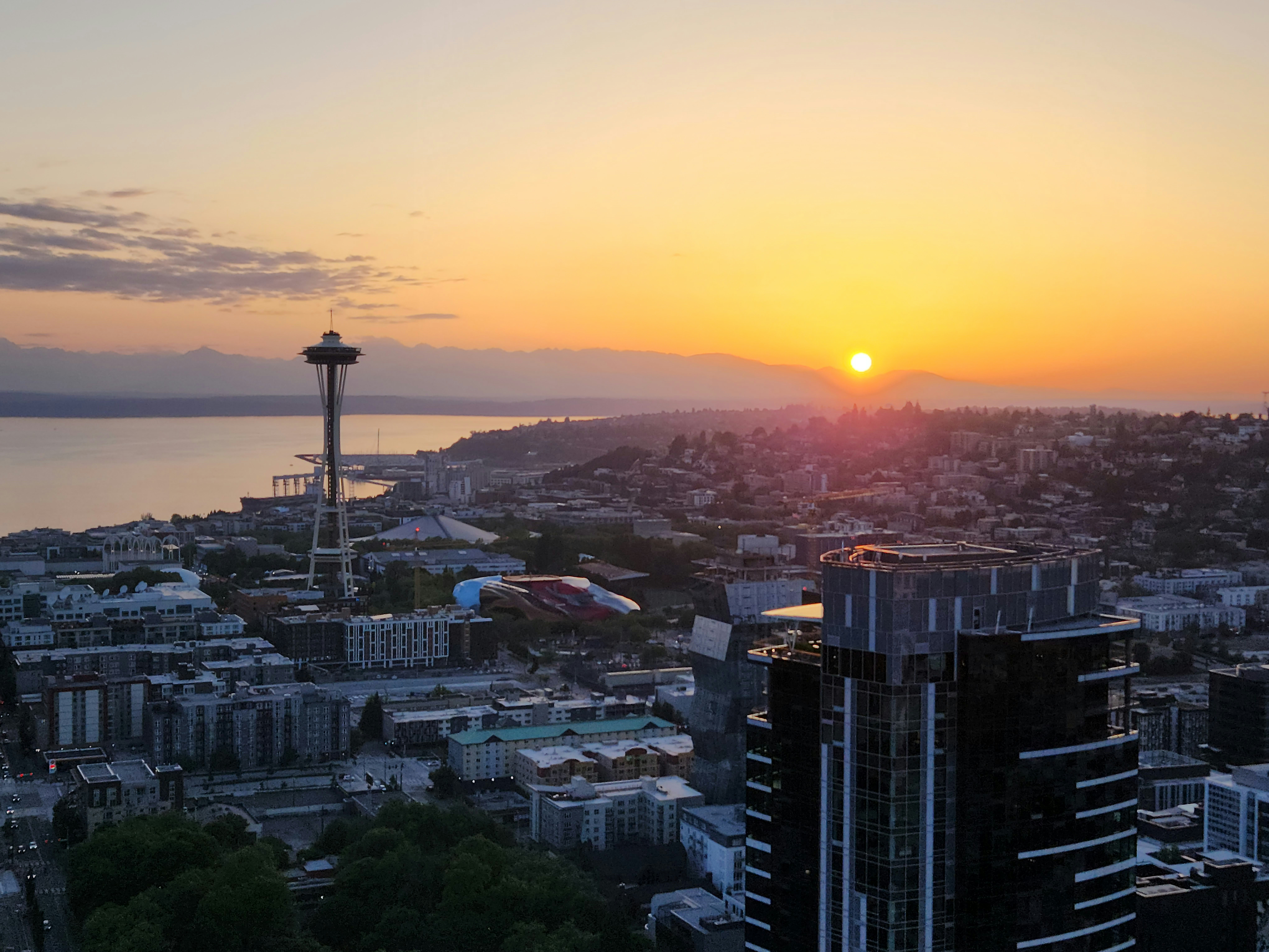 the sun is setting over a city with tall buildings, As the sun begins to set on the horizon, casting a warm golden glow over the city of Seattle, the Space Needle emerges as a magnificent silhouette against the breathtaking backdrop of the Olympic Mountains.
