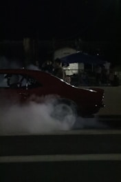 A car is performing a burnout, emitting thick smoke from its tires. The scene takes place at night with dim lighting. People are visible in the background, watching the event.