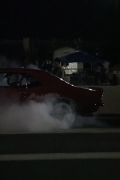 A car is performing a burnout, emitting thick smoke from its tires. The scene takes place at night with dim lighting. People are visible in the background, watching the event.