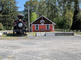 A vintage steam locomotive is displayed in front of a small, rustic train station building labeled 'ELBE'. The building is painted in a shade of coral with white trim and a decorative sign above the entrance. Surrounding the scene are lush green trees and a gravel area in the foreground.